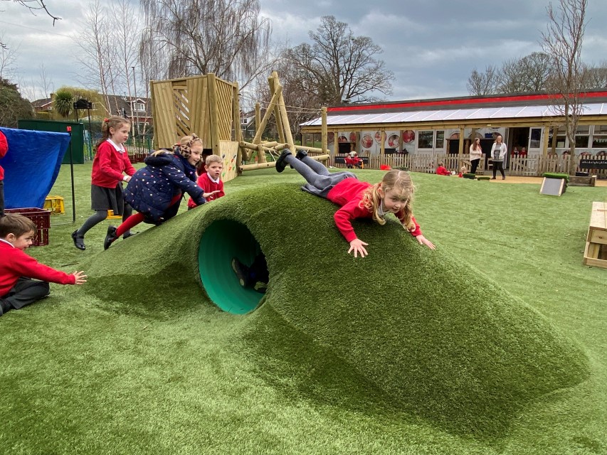 A little girl is sliding down an artificial grass hill with a tunnel in the middle. Another group of children are following close behind.