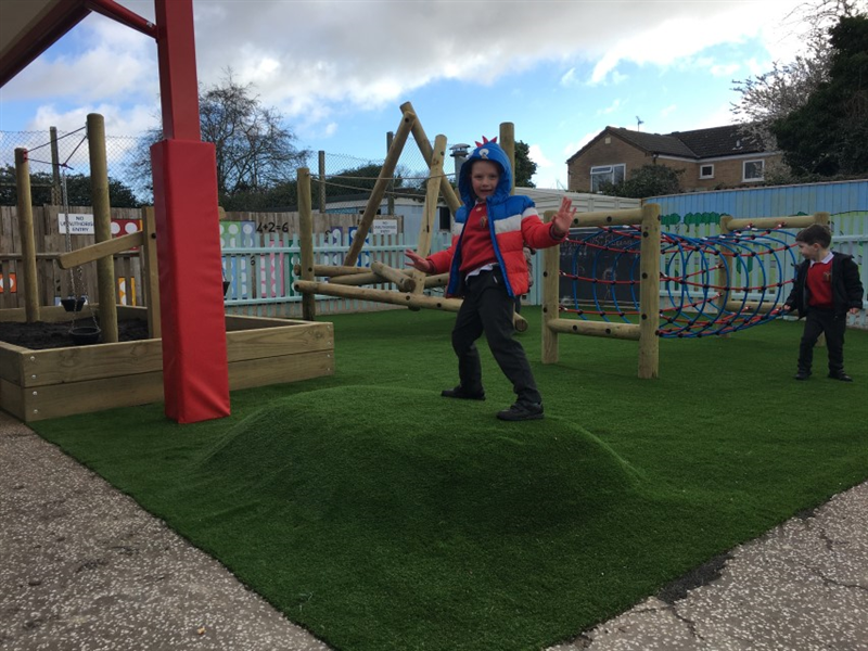 one boy stood on a playground surface mound wearing a red, white and blue coat posing for the camera with more playground equipment in the background including a net tunnel, climbing frame and rope and pulley.  