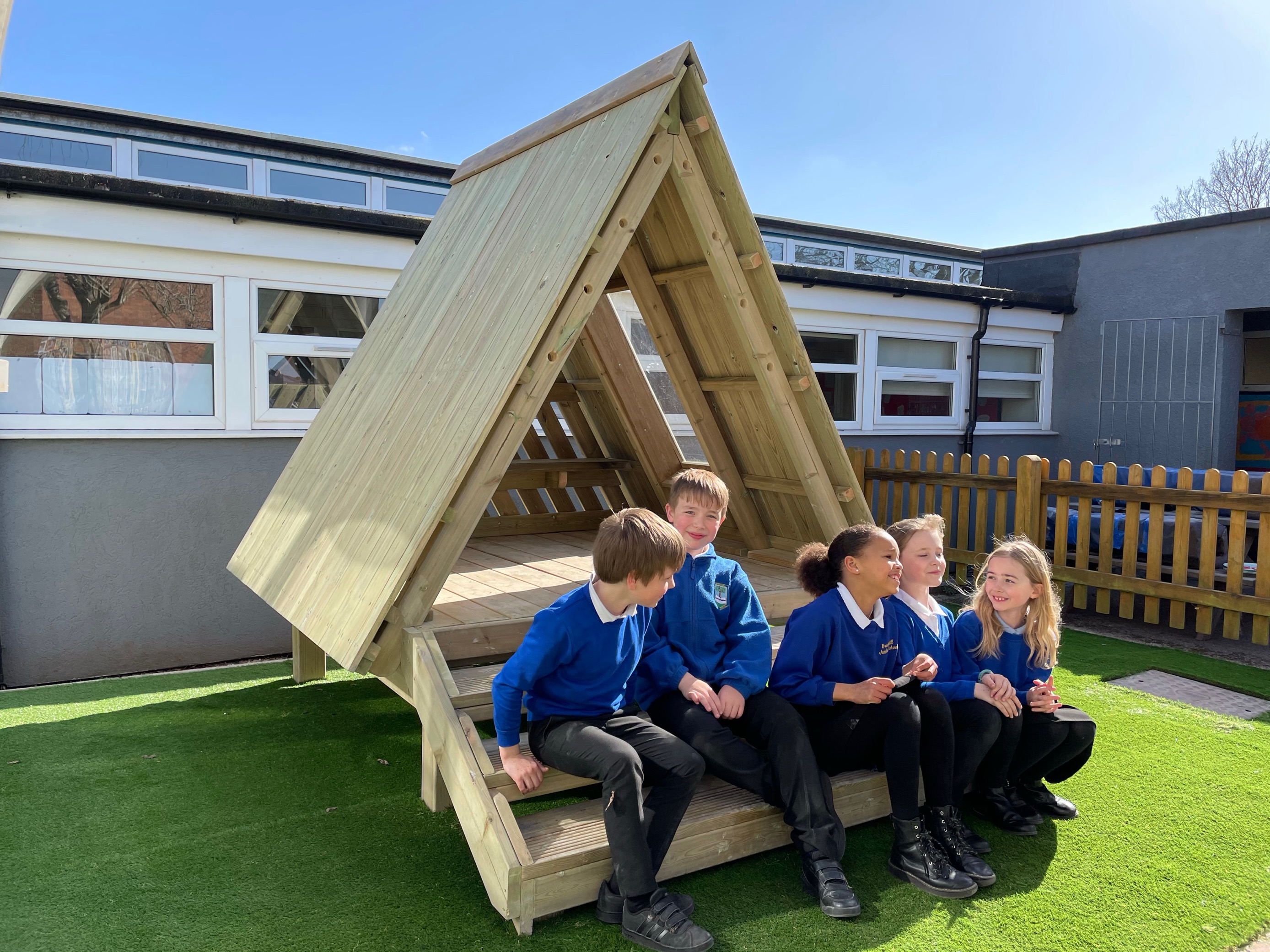 A group of children are sat beneath a wooden tee-pee and are talking and smiling with each other.