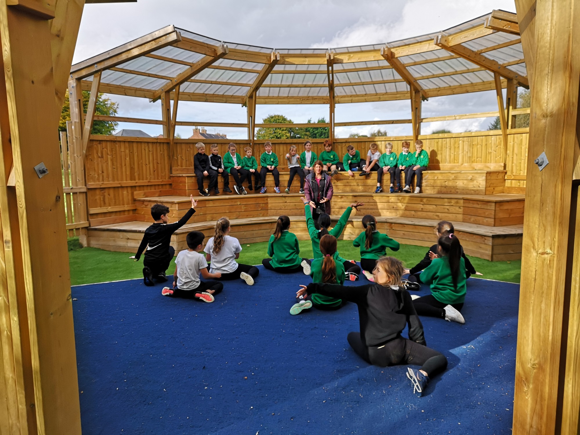 A wooden structure with 3 layers of seating. The top layer of seats has a lot of children sat on it, as they look down towards the stage, with other children sat on the stage facing a teacher.