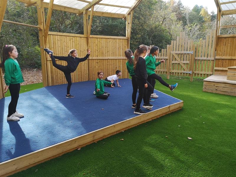 children perform yoga on a pentagon play performance stage which is timber with blue safer turf