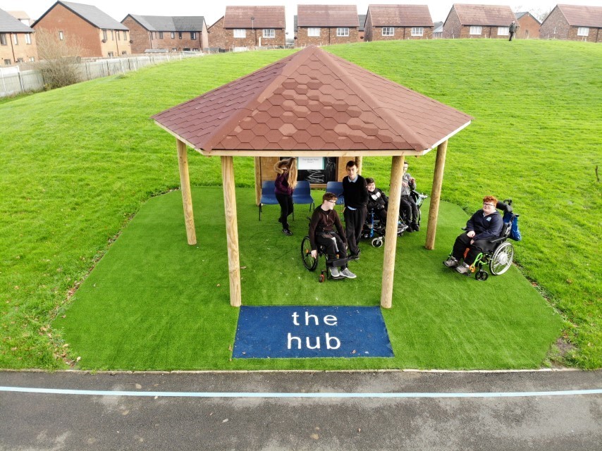 A group of children are stood and sat underneath a freestanding canopy, as "the hub" can be seen written beneath it.
