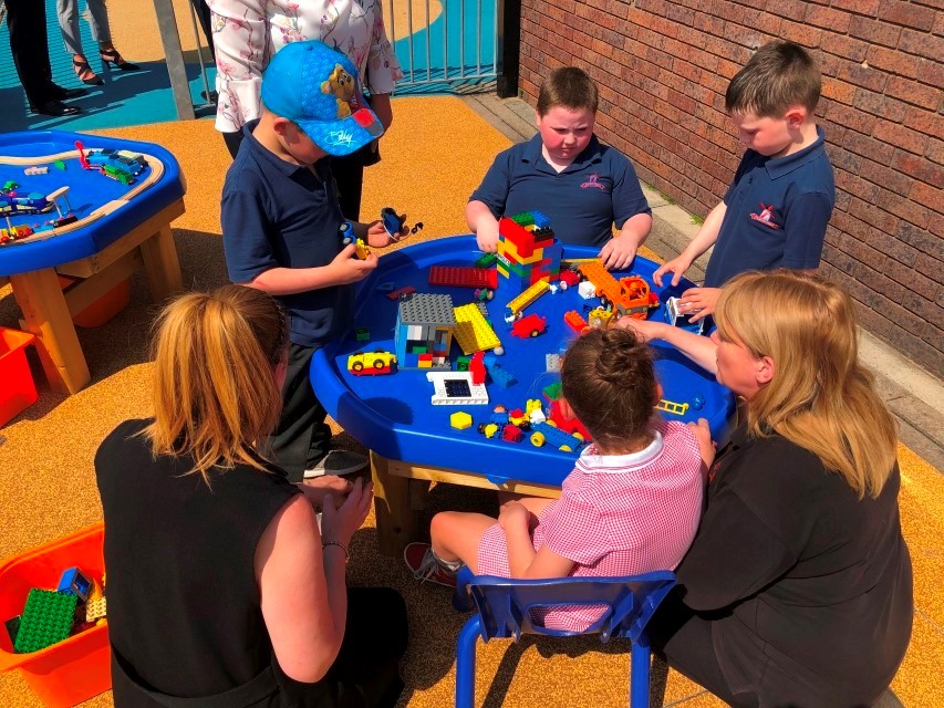 A group of children are playing with toys that are inside a tuff tray. Two teachers are crouching next to a little girl and are playing with the group.