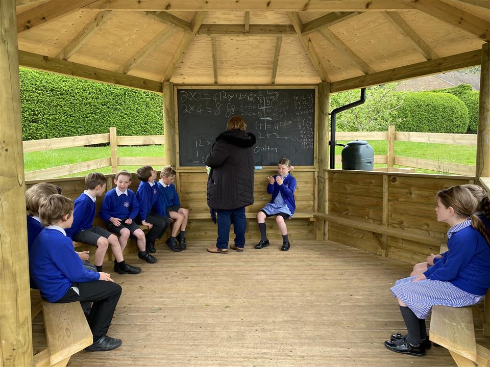 A teacher is delivering a lesson to her class of children in an outdoor classroom gazebo