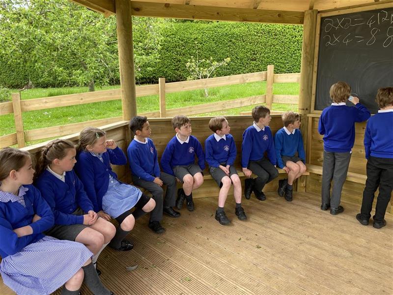 children in blue school uniform sit inside an outdoor classroom on a bench looking at the chalkboard