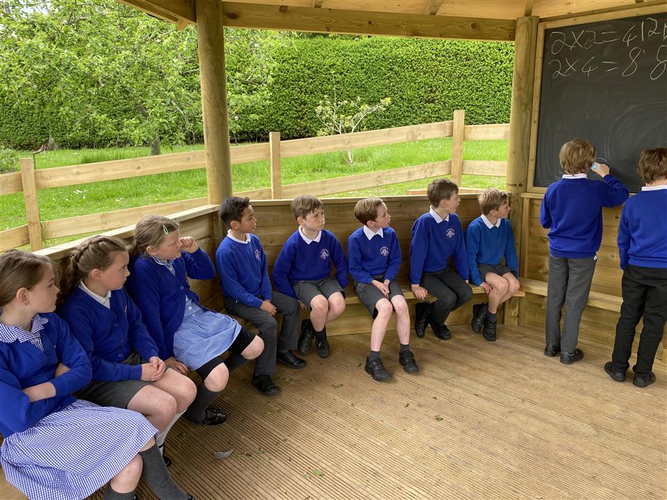 A group of school children are sat on benches within their outdoor classroom and are paying attention to the blackboard.