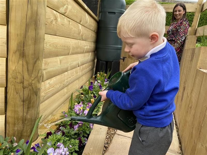  child in a blue school jumper holds a green wtering can and waters the plants in the flower bed