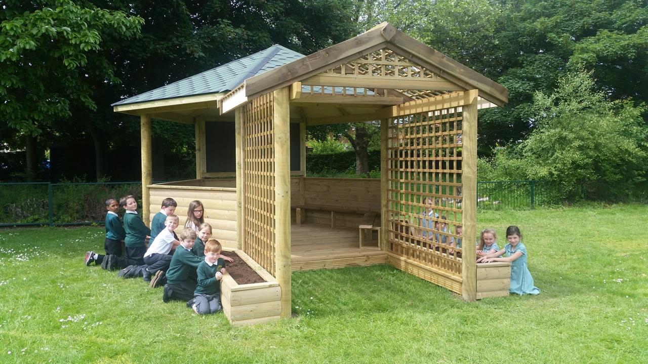 A group of children are kneeling beside their outdoor classroom as they are messing with the planters.