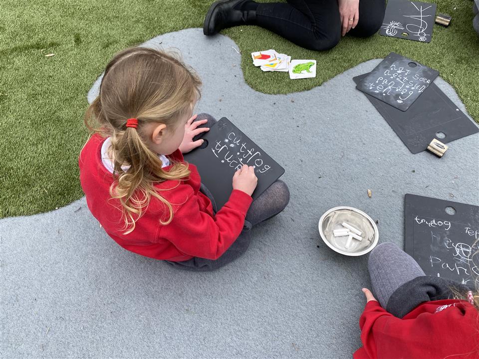 A little girl is writing on a small chalkboard that she has in her lap. She is writing different words on the board as she draws lines across the board. A teacher and another student are sat next to her.