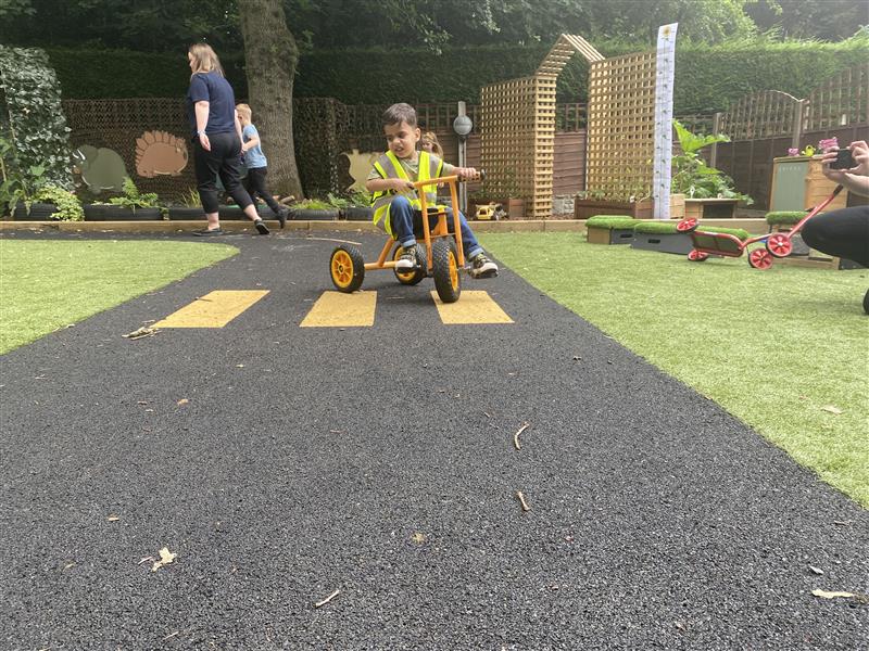 a child in a hi vi vest scoots around on the wetpour roadway towards the camera