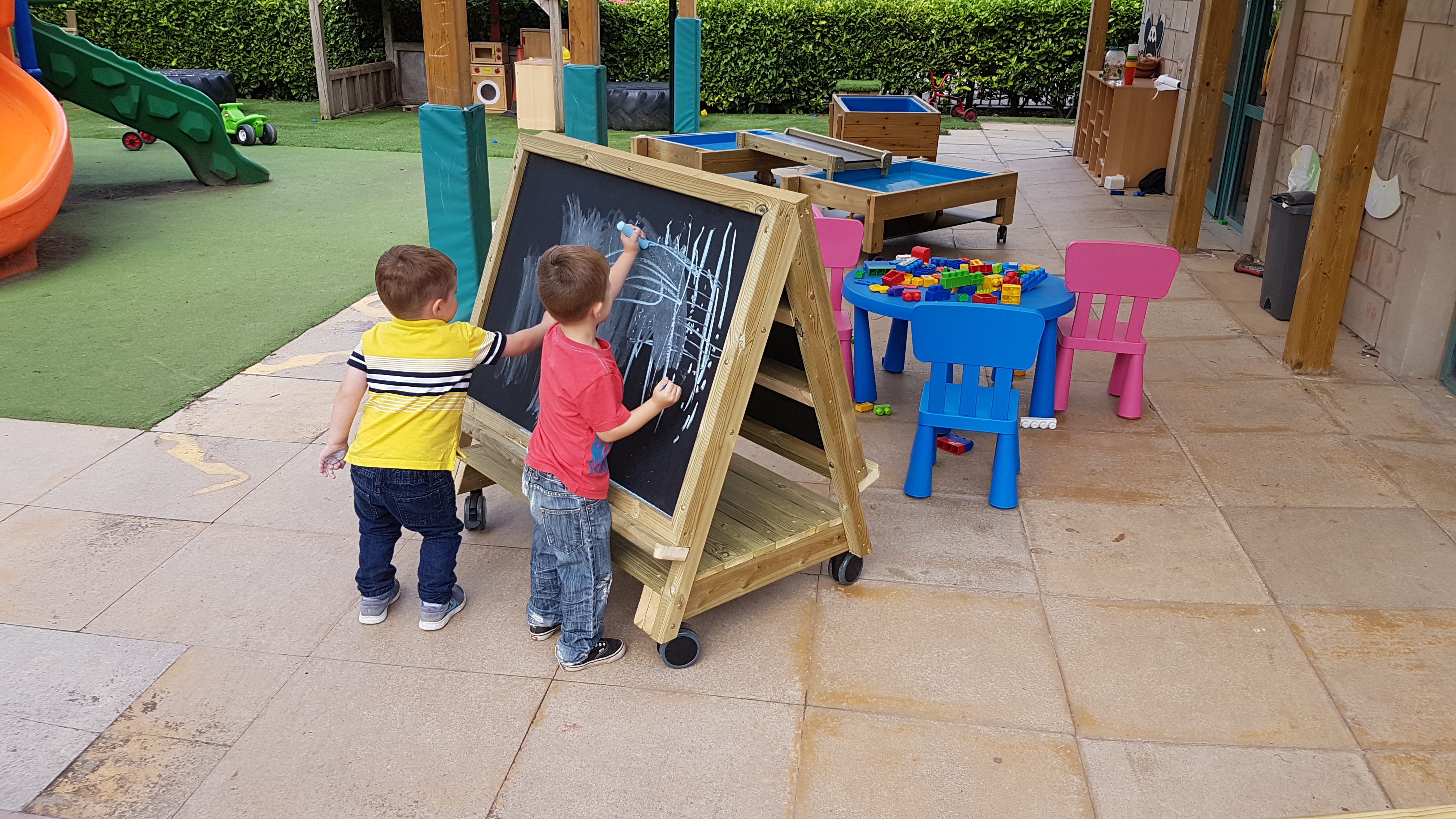 Two little boys are drawing chalk on a movable art easel, which has been placed outside of the classroom.