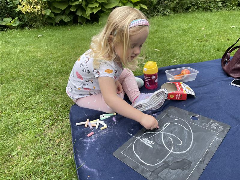 a little girl with blonde hair sits cross legged on the floor and draws on the whiteboard with white chalk
