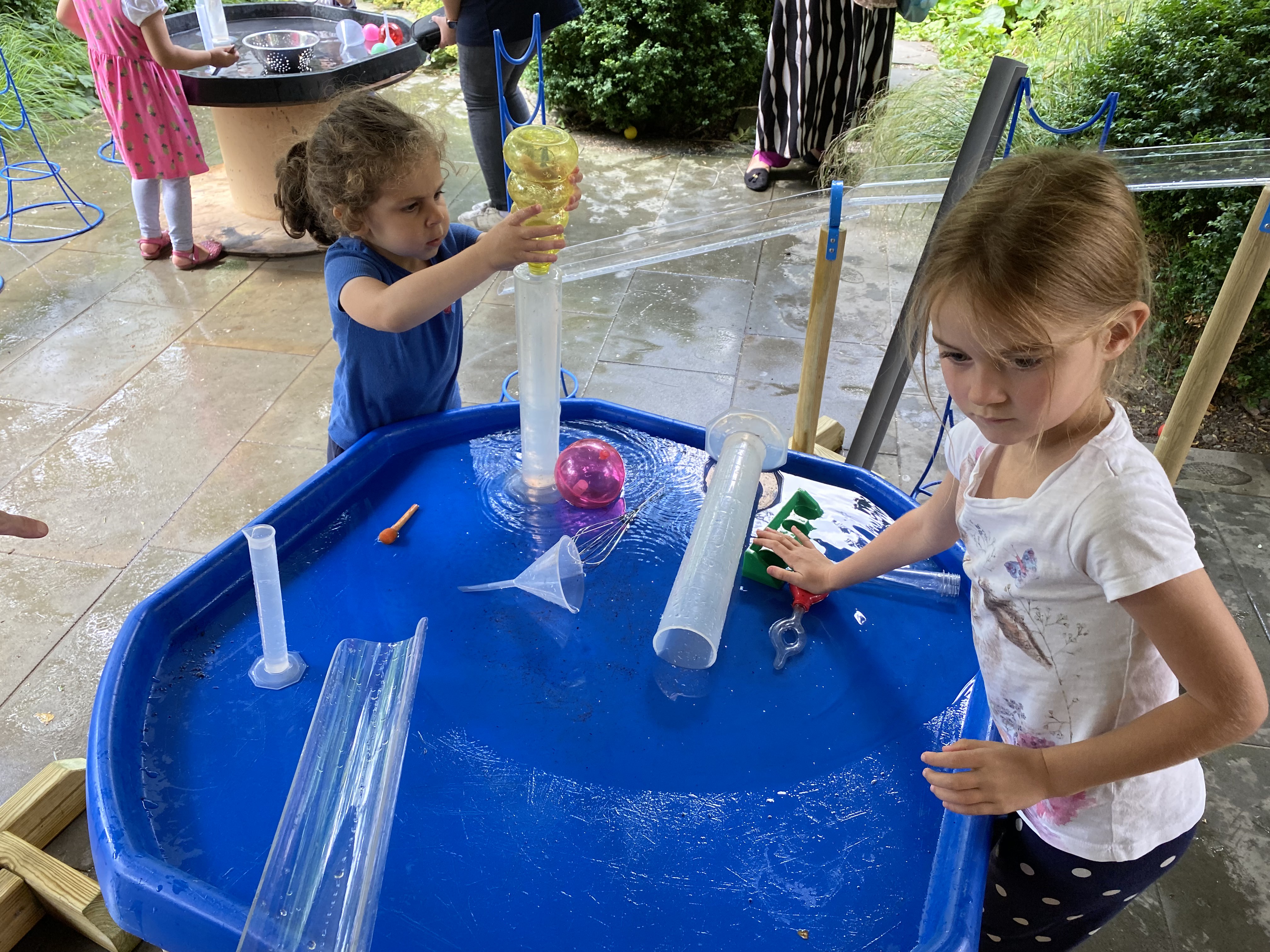 Children are playing with a water table that has been placed outside. The children are splashing around the water and using a variety of water play tools.