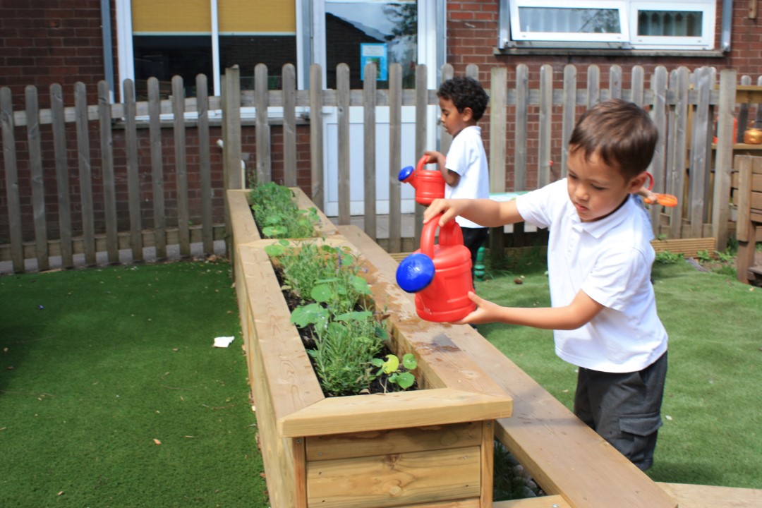 2 children are holding small, red watering cans as they water the flowers that are planted in wooden planters. These planters are raised, allowing the children to stay stood up as they water the plants.