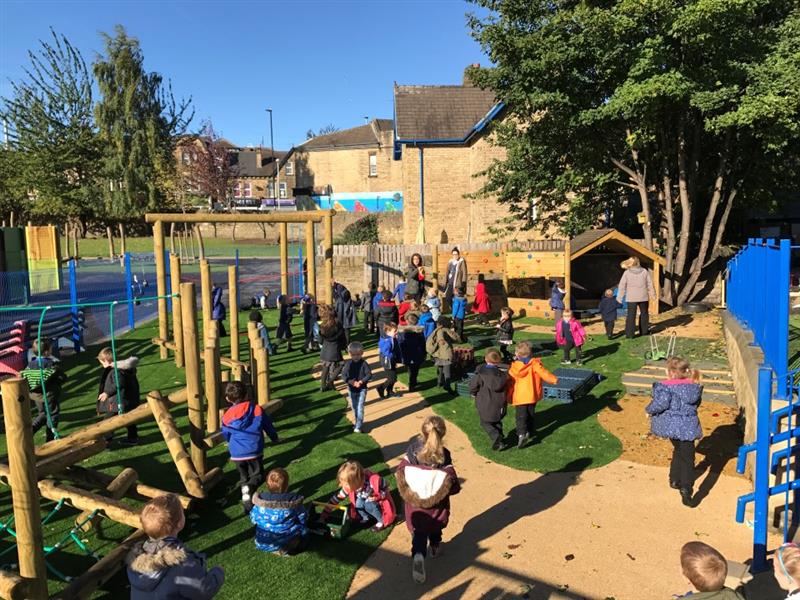 a busy and hectic eyfs area with beige wetpour and artificial grass surfacing, children in blue school uniform and coats run around playing on the ytrim trail which consists of steppig logs, a rope traverse, and a climbing wall, there is alsoa giant play house with chalkboard and benches