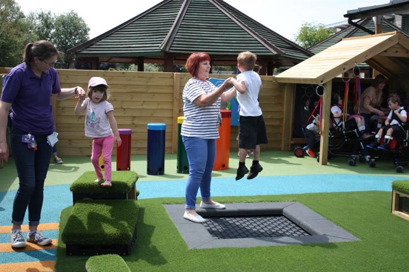 a teacher helps a child in school uniform bounce on the sunken trampoline which is surrounded by our luscious green artificial grass, a child next to him climbs along the get set, go clocks with the helping hand of another teacher on a sunny day