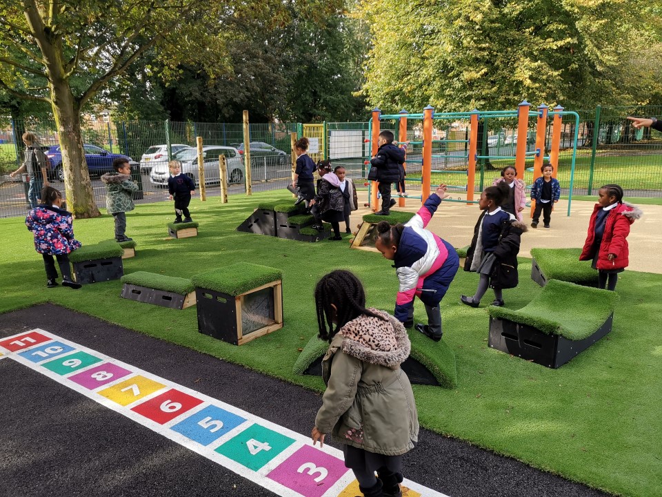 A large group of children are playing on freestanding play equipment and are playing together. The equipment has been set up on synthetic turf and is next to a thermoplastic hopscotch