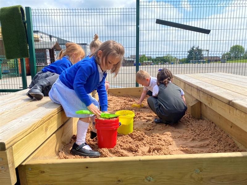 a little girl in blue school uniform crouches as she scoops sand into a red bucket using a green spade as her classmates play in the sand behind her
