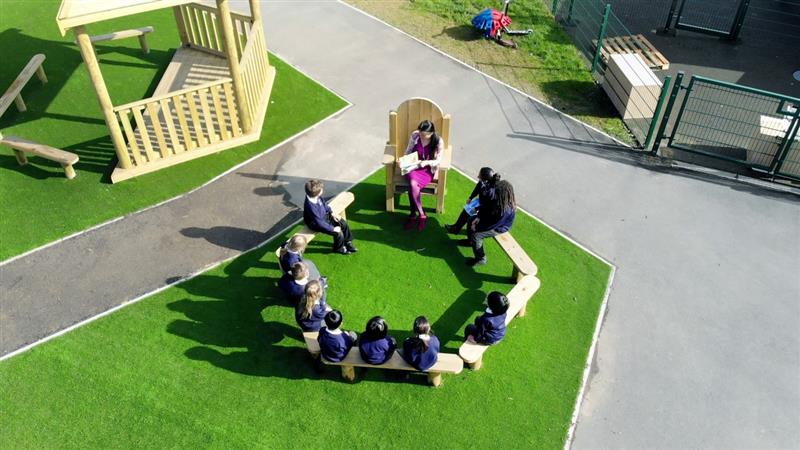 children seated on benches in a circle whilst a teacher reads a story to them as they sit and listen
