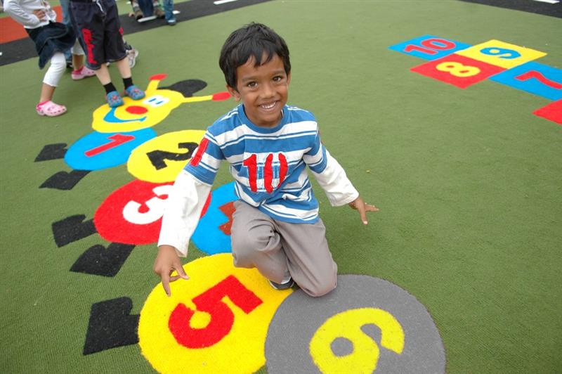 a child in a blue striped top smiles at the camera as he kneels on our brightly coloured caterpillar playground marking on our green artificial grass