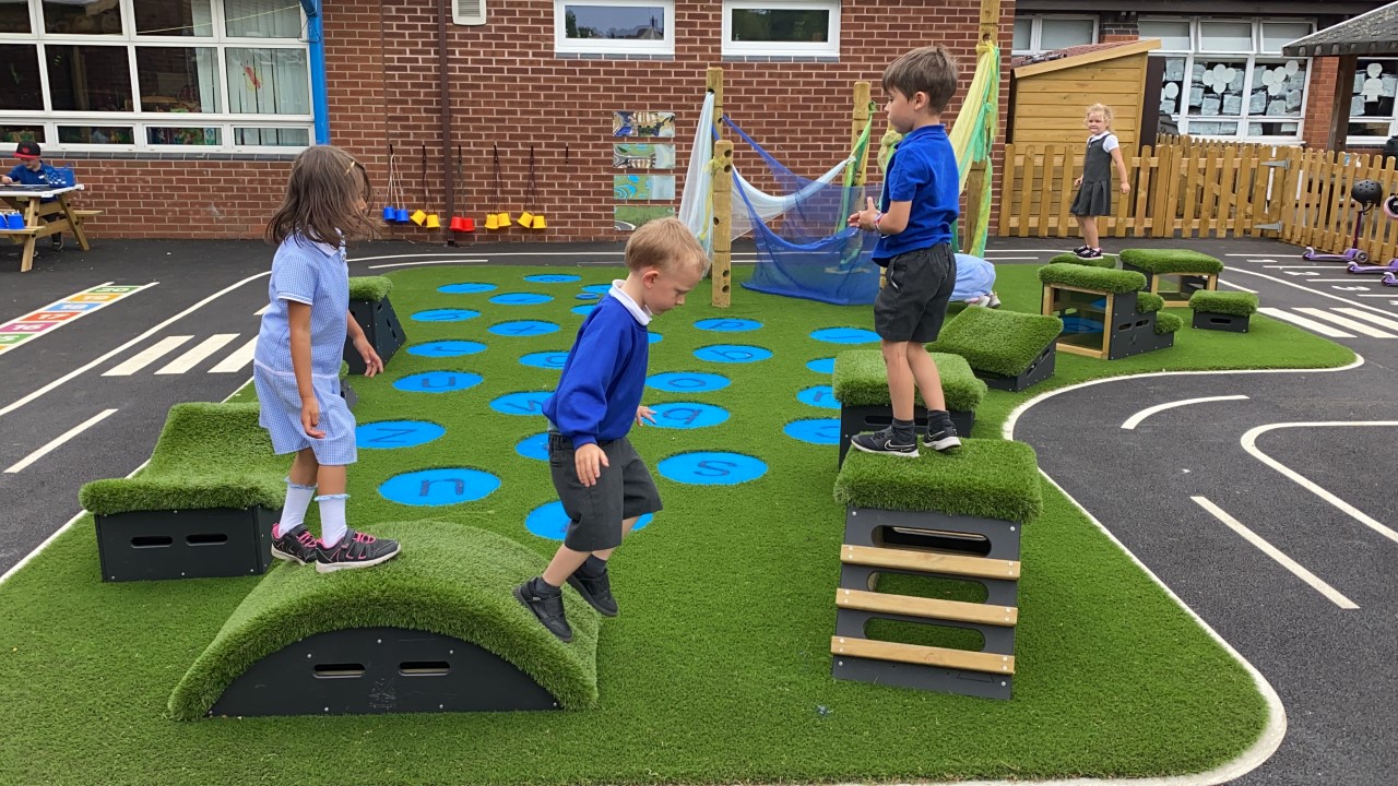 A group of children are playing on freestanding play equipment. The equipment has been placed on artificial grass and has a wetpour road going around the area.