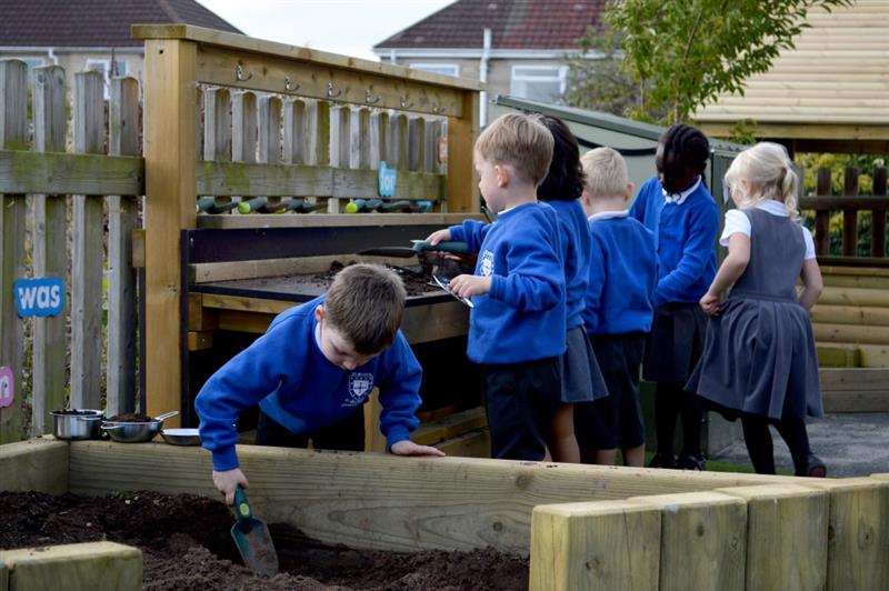 a view of a child in a blue school uniform digging into a dig pit ith a shovel whilst his classmates play with a mud kitchen behind him