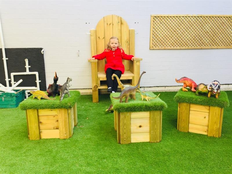 a child sits on the story telling chair and looks at all different toy dinosaurs sat on artificial grass toys