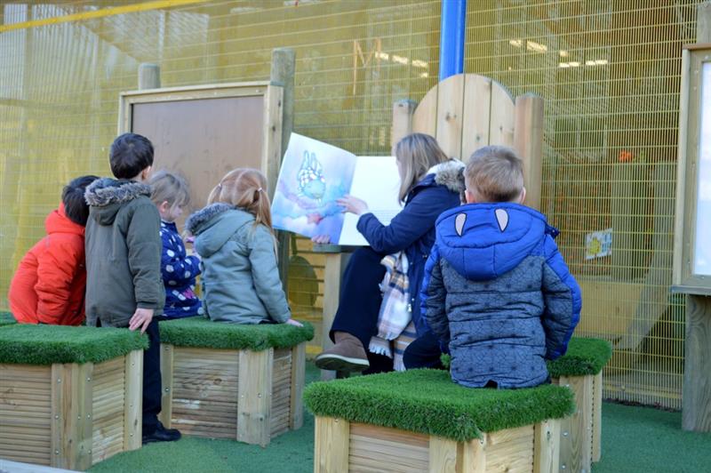 little children in winter coats sit on artificial grass topped seats in front of a story-telling circle  whilst being read a story book by a teacher also in a winter coat