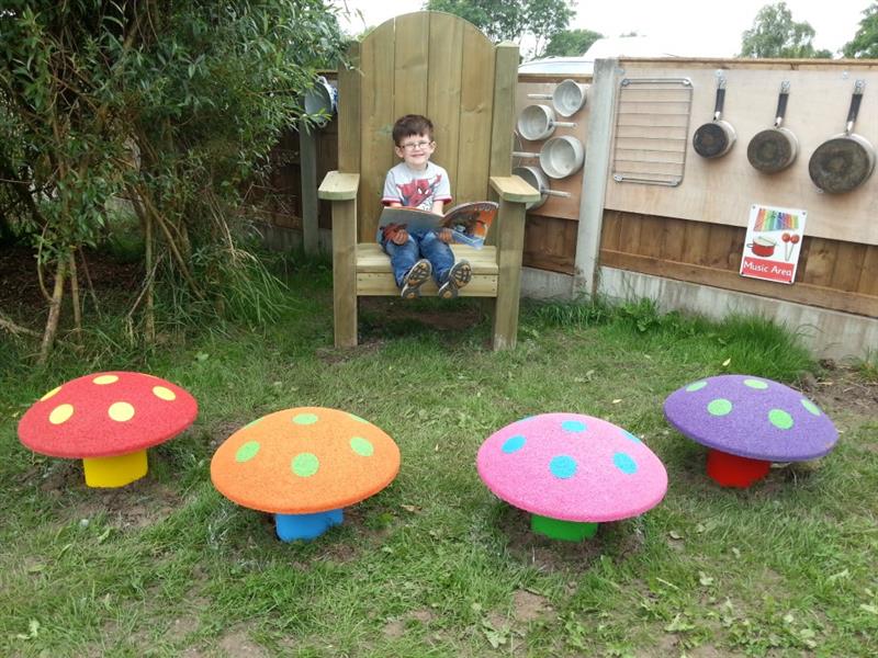 a little boy sits in a large story telling chair with his feet up reading a book with colourful mushroom seats gathered around in front of him in a semi-circle