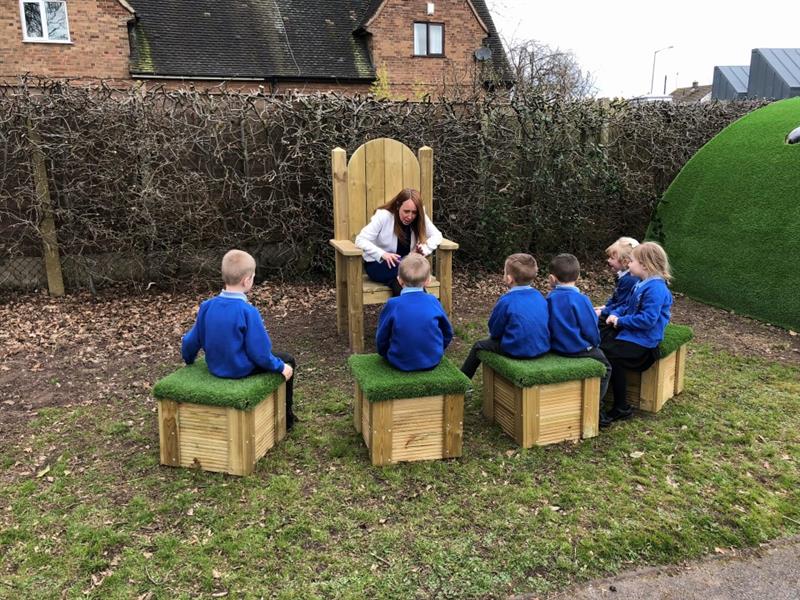 children sit in a semi circle on the artificial grass topped seats in front of their teacher who sits in the timber storytelling circle telling the children a story whilst wearing a white jacket