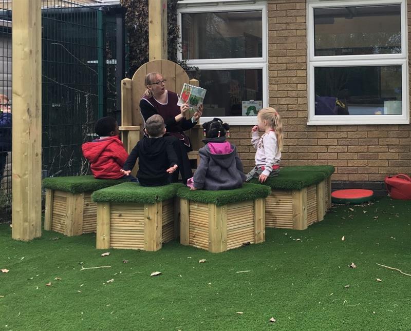 a lady sits on a story telling chair and reads a picture book to the children sitting on grass topped seats