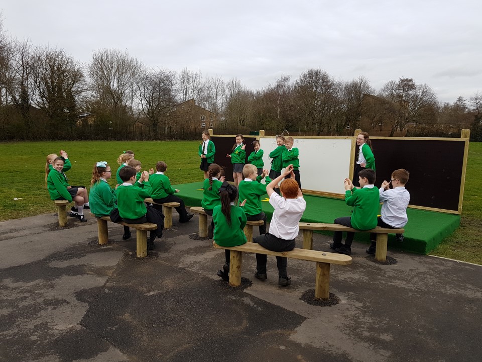 A group of children are stood on an outdoor stage and are posing for the audience. A large group of children are sat on wooden benches opposite and are clapping for the performance. Behind the stage is a whiteboard with a chalkboard on either side. The stage is covered in green saferturf.