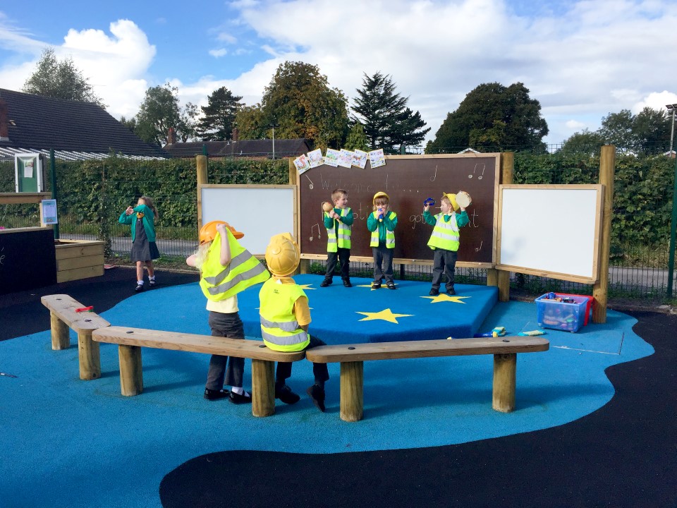 3 children are stood on an outdoor stage as they wear hi-vis vests and yellow hardhats. 2 children are sat on benches opposite the stage, wearing the same hi-vis and hardhat as the performers. A little girl is stood to the right of the stage.