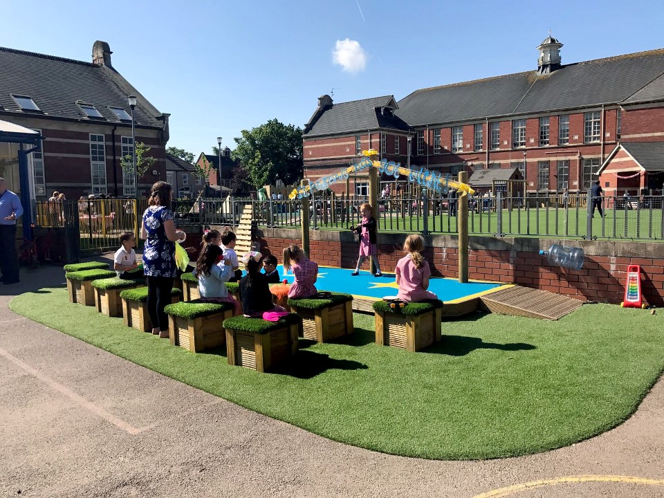 A girl is performing on a stage in front of a class of children. The stage is outdoors, with a blue and yellow surface. The children watching are sat on wooden boxes with artificial grass tops. The teacher is stood up at the back, watching the child perform.