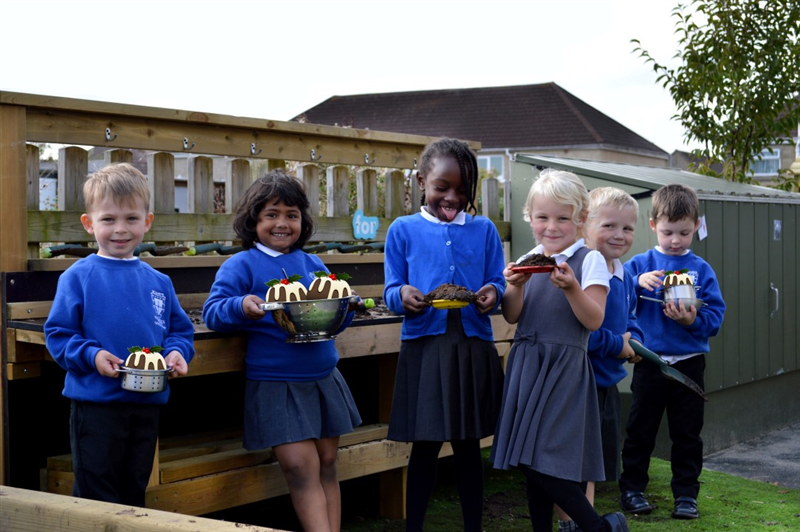 six children stand in front of a mud kitchen and gather around holding silver bowls with christmas puds in it