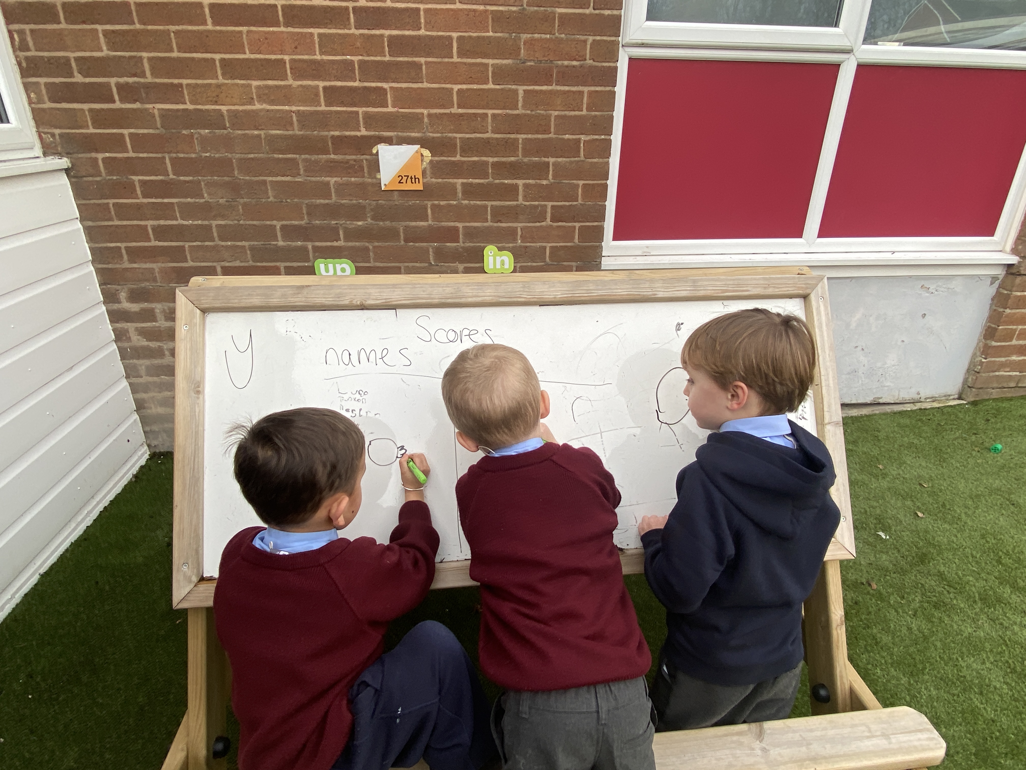 A group of three children are stood around a whiteboard easel, each holding a black whiteboard marker. The children are writing on the whiteboard easel. The whiteboard is slightly angled, allowing the children to get a better position with writing.