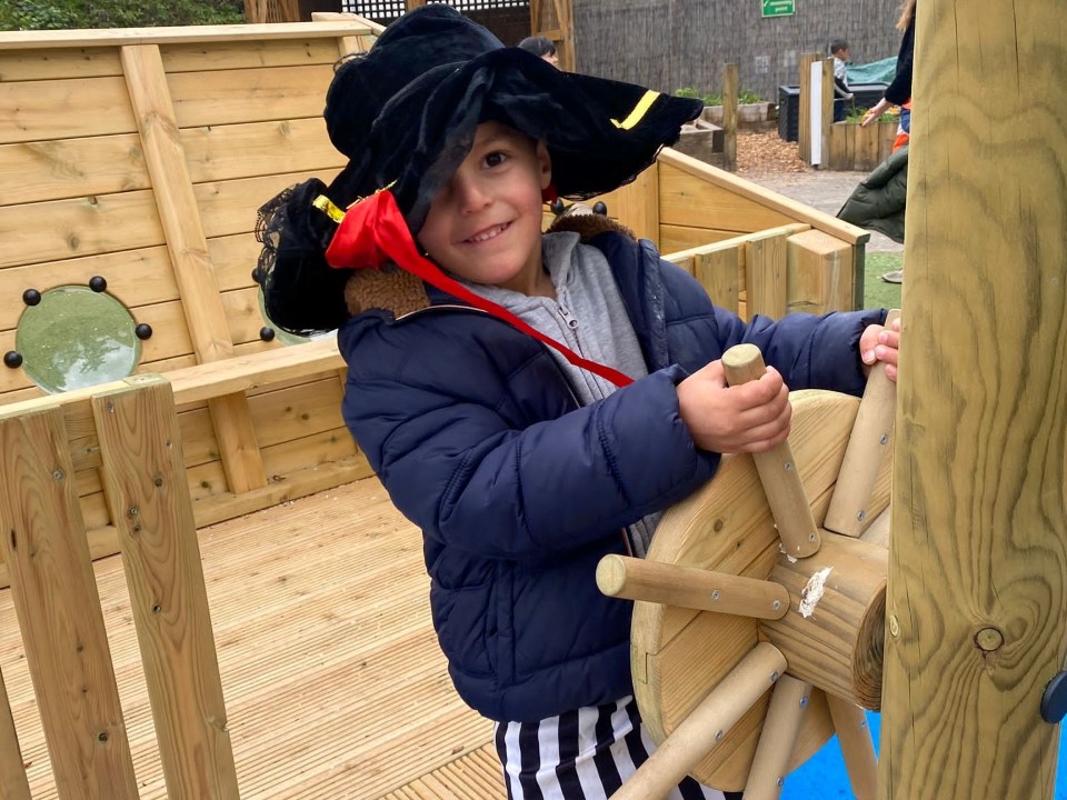 A little boy is dressed as a pirate, with a fake parrot on his shoulder. He is holding onto a wooden pirate ship wheel as he looks into the camera.