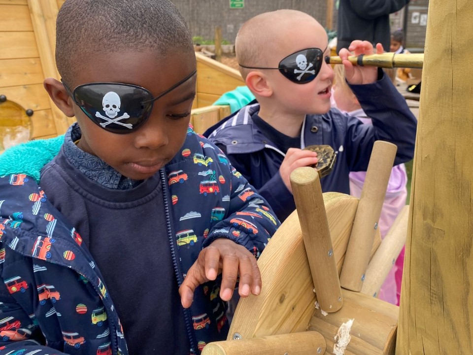 2 children interacting with the low level outdoor play ship, which was created by Pentagon Play. The two children are wearing eye-patches, with one child holding a telescope. They are both playing with the steering wheel.as they engage in pretend play.