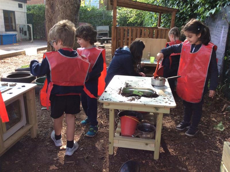 children stand in messy play aprons around the mud kitchen