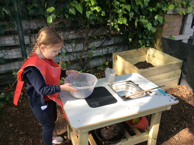a little girl stands with a bowl and mixes things together at the mud kitchen island