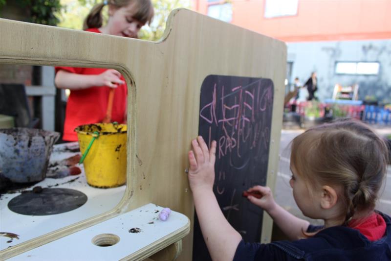 a little girl kneels and draws on the chalkboard on the back of the mud kitchen