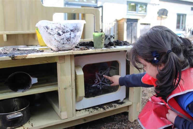 a little girl kneels down in front of the mud kitchen and points into the oven