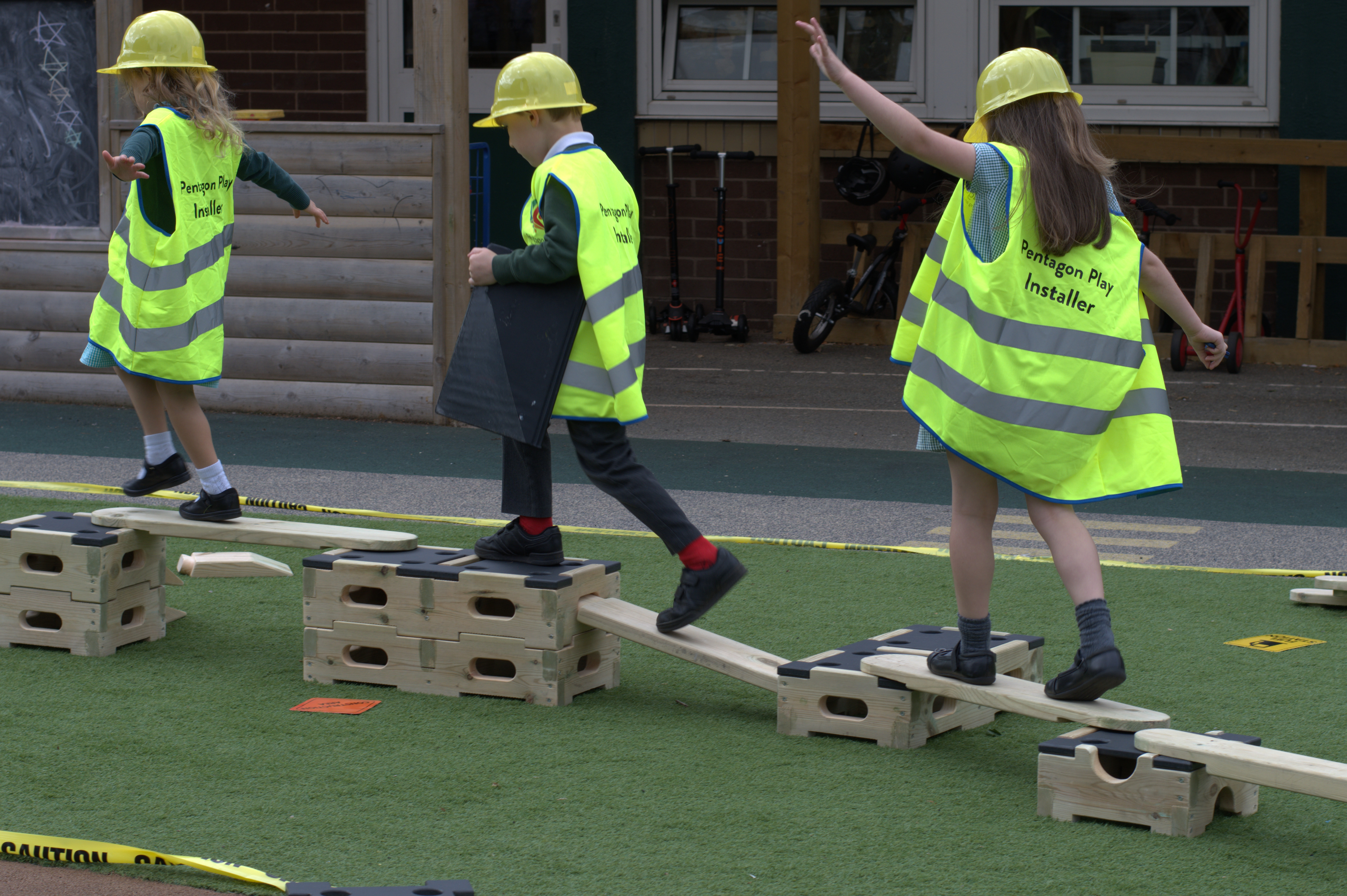 Three children are playing on a play builder set, wearing hi-vis jackets and hard hats. They are balancing across wooden planks in a single file line.