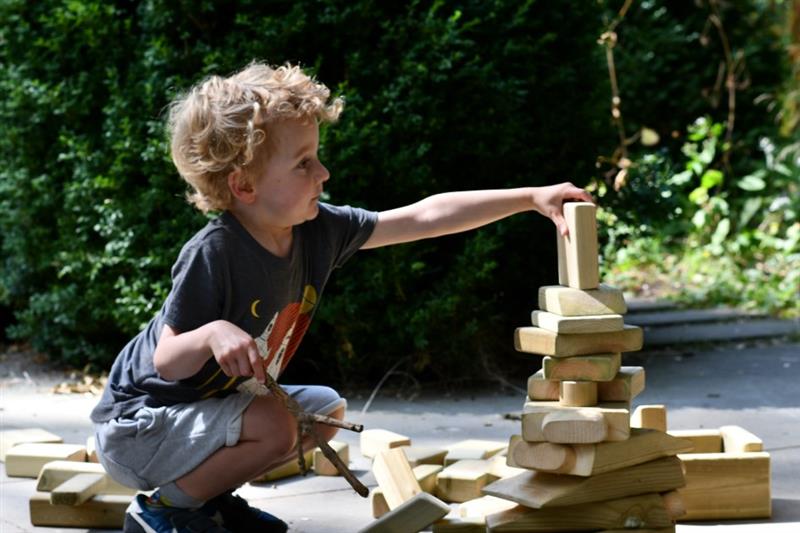 a little boy sits on the floor of a playground and builds a tower out of the wooden blocks that come in the construction skip