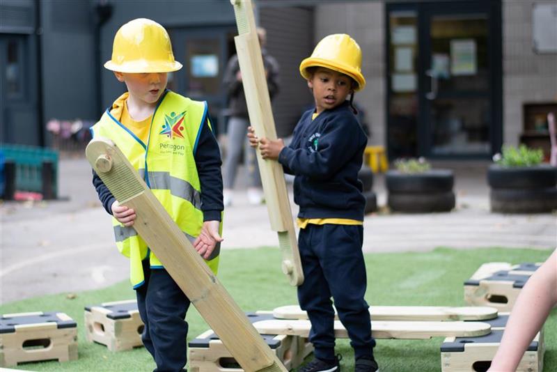 two children, dressed in hi vis vests and pretend hard hats carry play builder planks around the playground