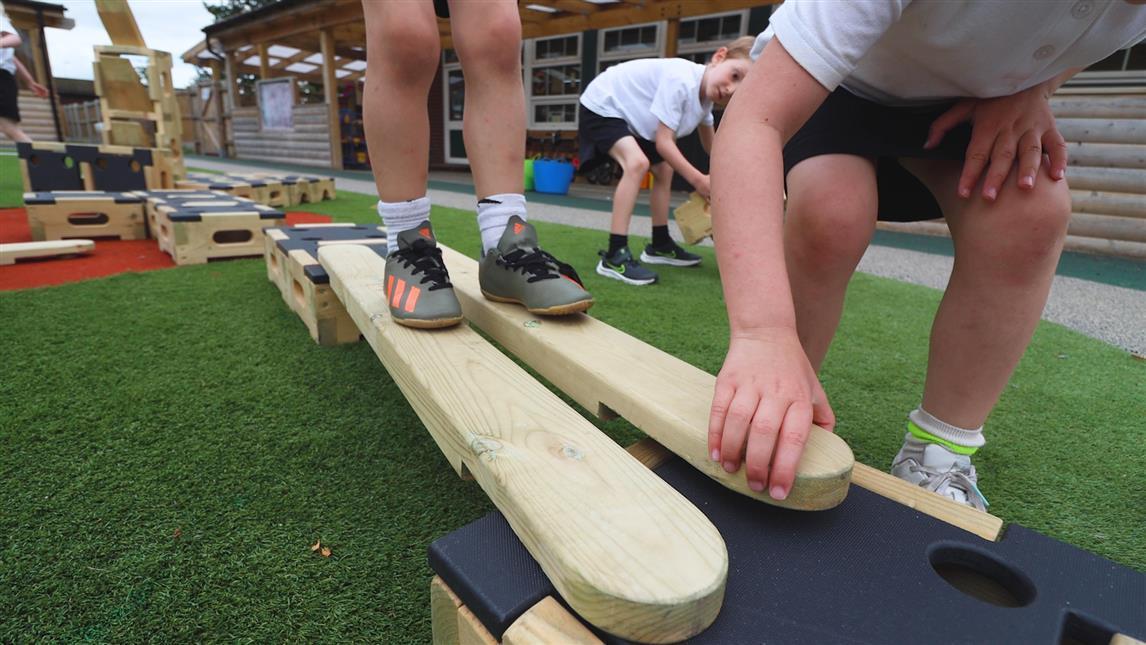 A child is connecting a plank to a play builder block, as another child balances across the plank.