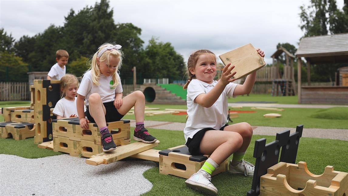 4 children are building an obstacle course with the play builder set. The girl at the front is pulling a funny face as she holds up a wooden block to the camera as the three other children are occupied with building paths and obstacles.