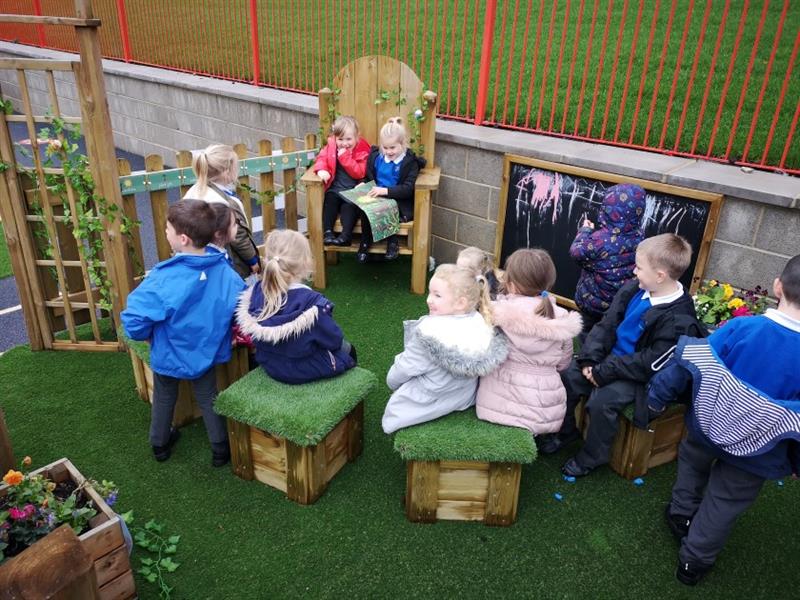 children sit on the storytelling chair and read a story together whilst a child draws on the chalkboard and others sit on the grasstopped seats