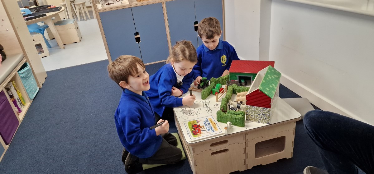 3 Children are sat at a whiteboard table and are drawing on it. Laid on the table is a farm with farm animals and a book. The children look very happy!