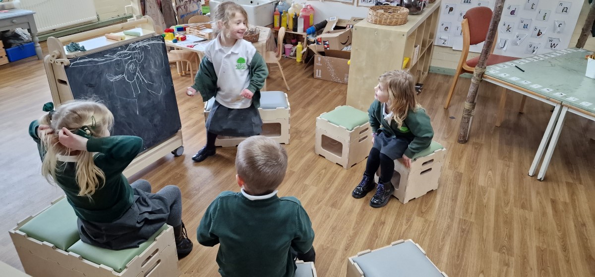 4 children are all sat around a chalkboard as one of the four children is stood up next to it and is presenting something to the rest. She is drawing an animal.